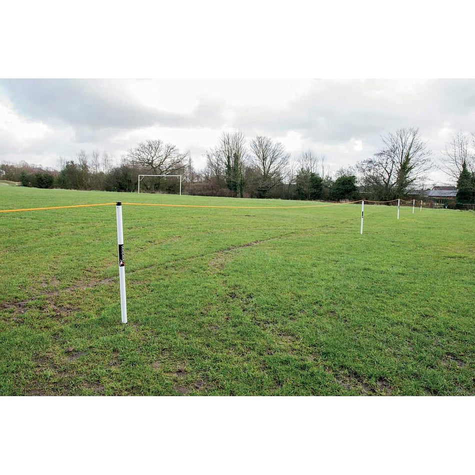 A grassy sports field with a soccer goal, bordered by Precision Training’s Precision Crowd Barrier—white posts and yellow tape—beneath cloudy skies, with a nearby tent and trees framing the scene.