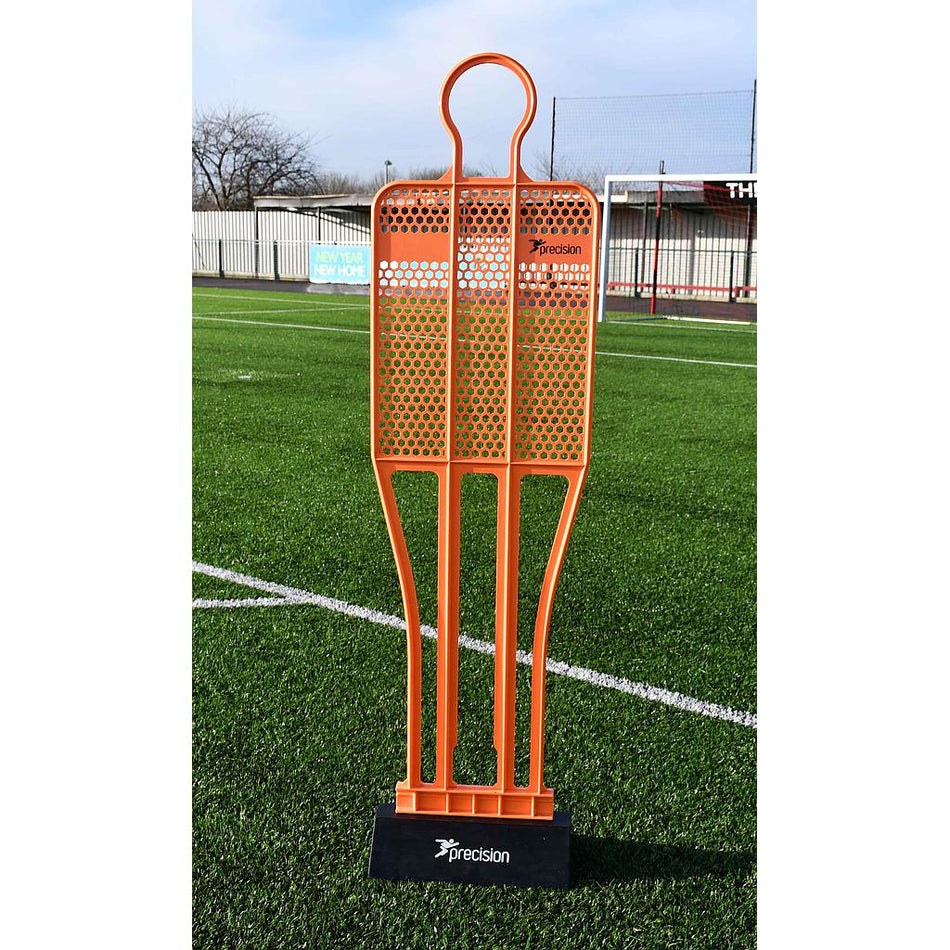 The Precision Training Pro HX Mannequin, in bright orange, stands upright on artificial grass—perfect for free kick practice. A sports facility with fencing and goalposts is visible in the background.