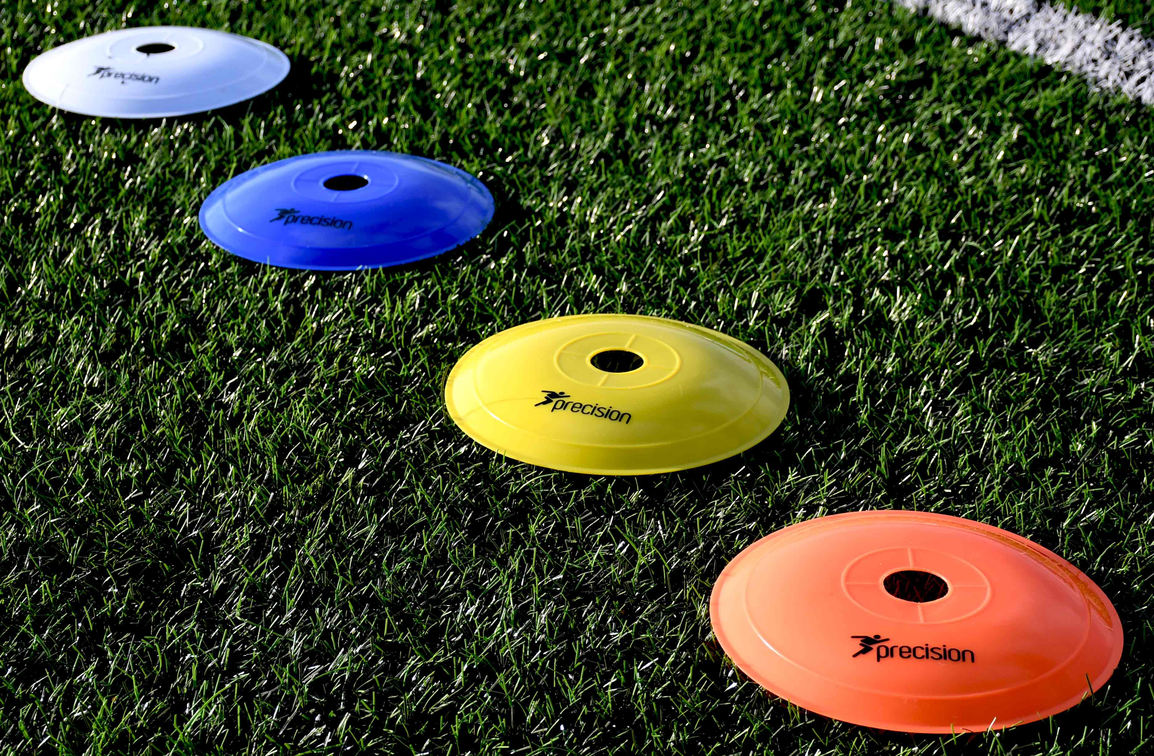 Four Precision Training Pro Near Flat Markers—white, blue, yellow, and orange—are lined up diagonally on green artificial turf. Each soccer training marker displays the brand name and logo. Set includes 40 markers.