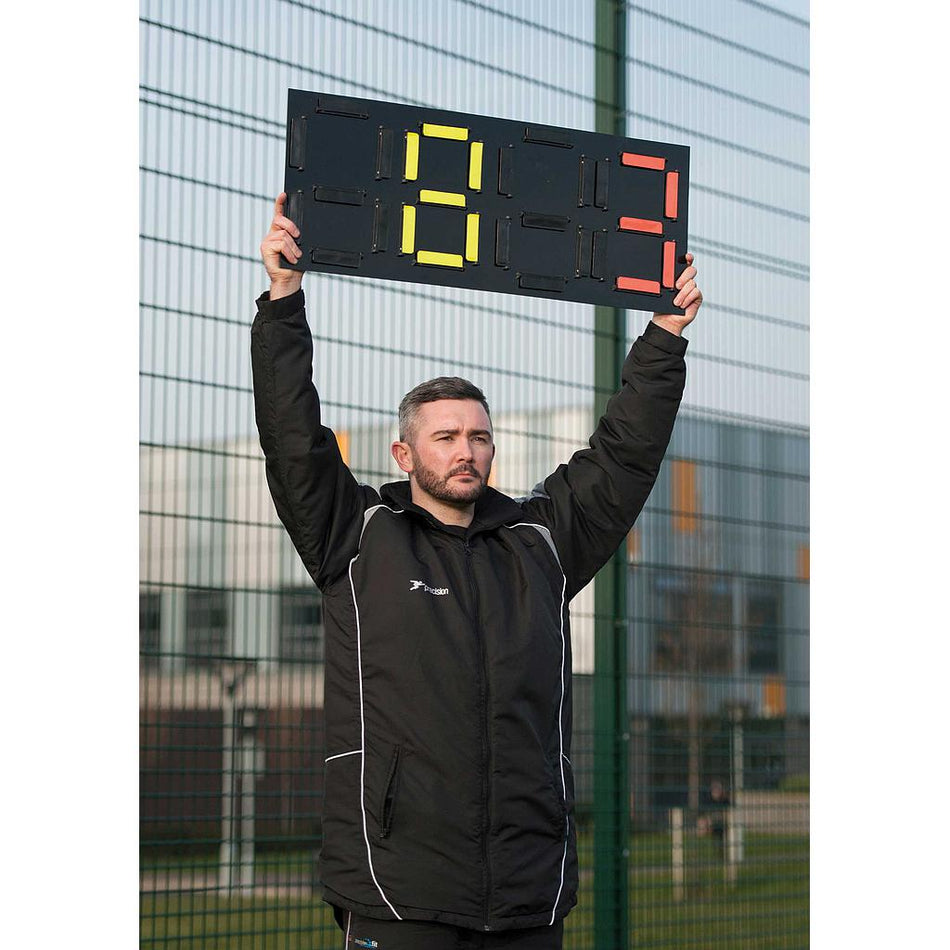 A man in a black jacket uses the Precision Training Precision Pro Substitutes Board, showing player numbers 6 in yellow and 3 in red, standing pitch side near a wire fence and modern buildings.