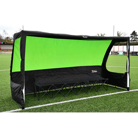 The Precision Pro Team Shelter by Precision Training is a black, quick-assembly pop-up sports shelter with a bright green back panel and foldable bench, shown on the sidelines of an outdoor artificial turf soccer field.