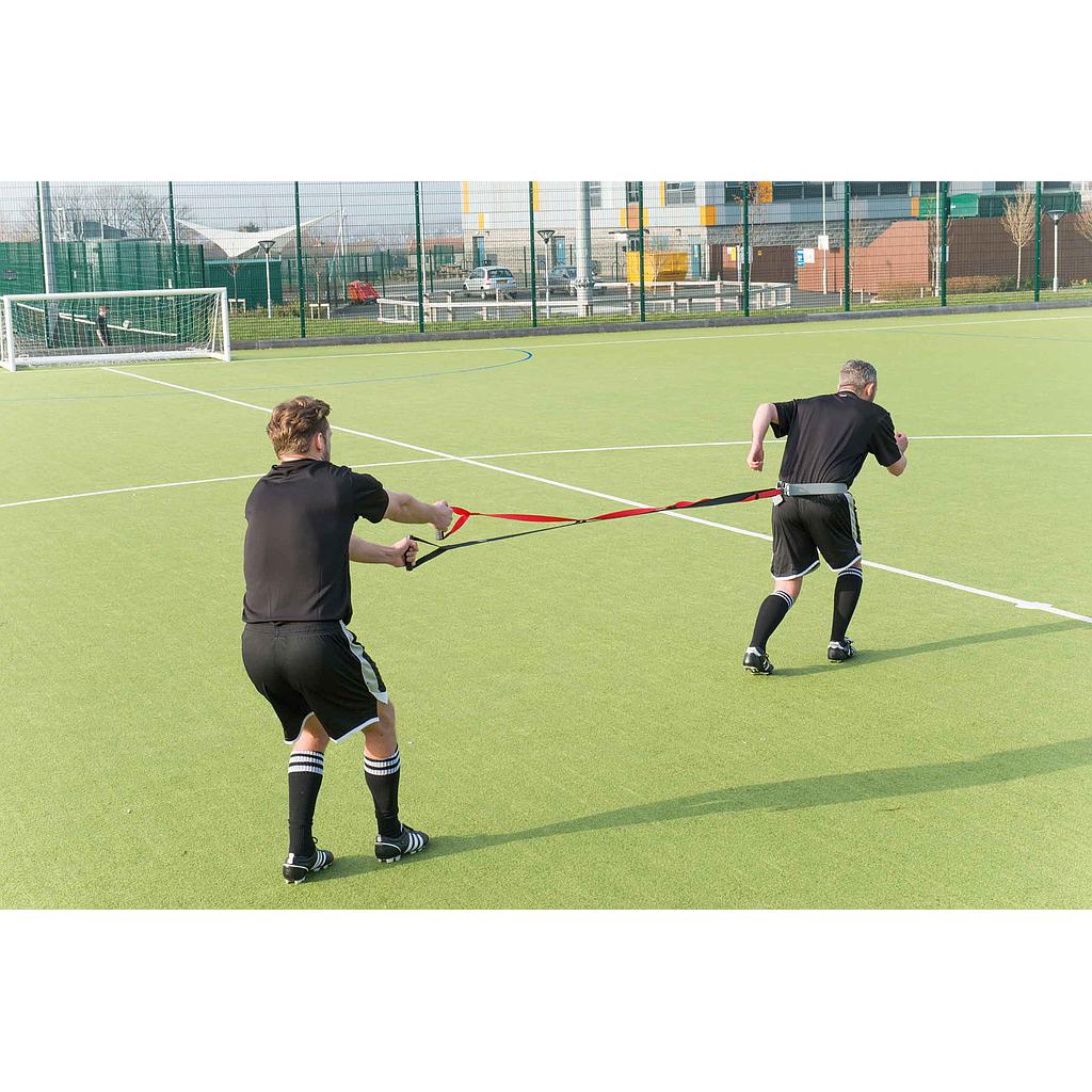 Two people in sportswear use the Precision Training Precision Pro Quick Release Belt for partner resistance training on an outdoor soccer field. One sprints forward as the other provides resistance. A goalpost and buildings are visible in the background.