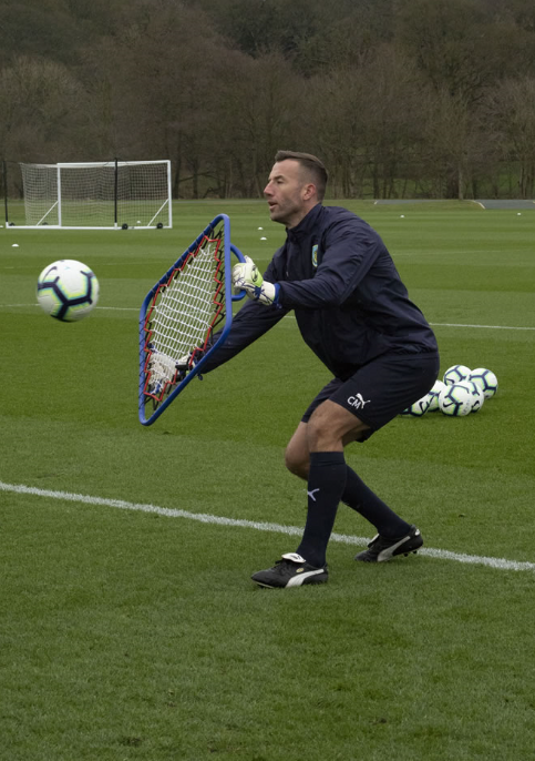 A soccer coach uses the Samba Sports Handheld Re-bounder to deflect balls during a goalkeeping session on grass, with multiple soccer balls and goalposts in the background.