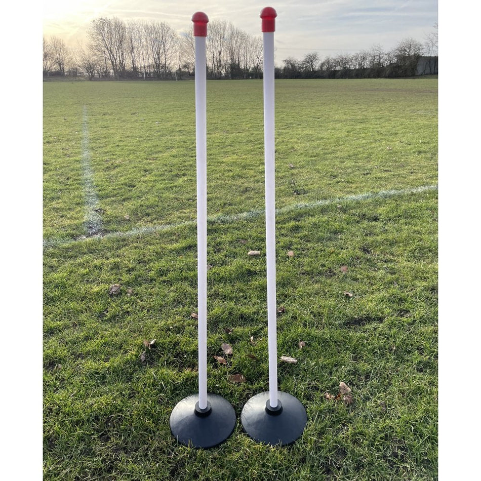 Sure Shot Wooden Rounders Posts by Sure Shot—two tall white posts with red safety caps and sturdy black bases—stand upright on grass, with trees and a pale sky in the background. Set includes 4 wooden posts.