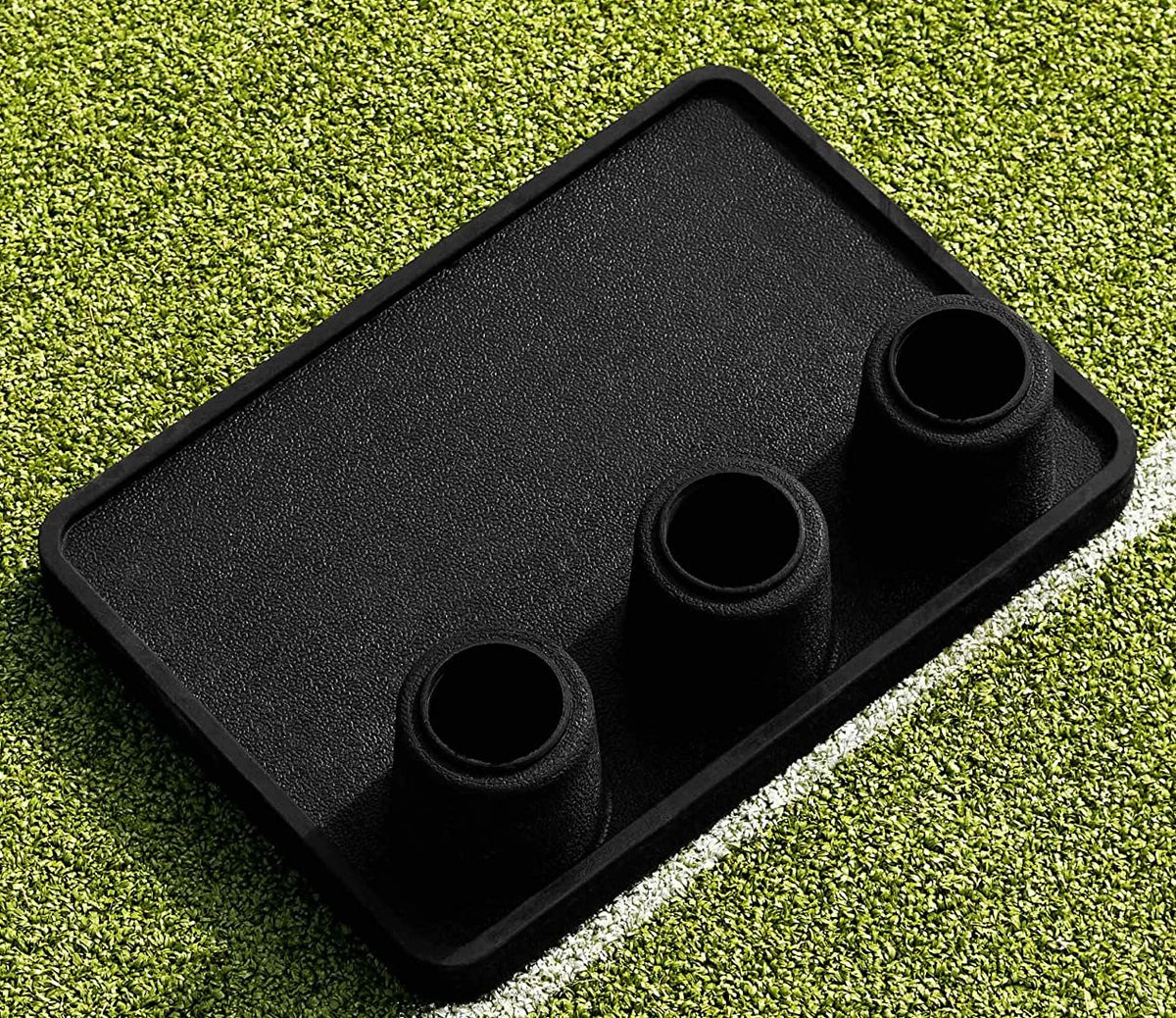 The Martin Berrill Sports Heavy Duty Rubber Flexi Stump Base (Black) with three cylindrical holders for plastic cricket stumps is shown on green artificial turf beside a white line.