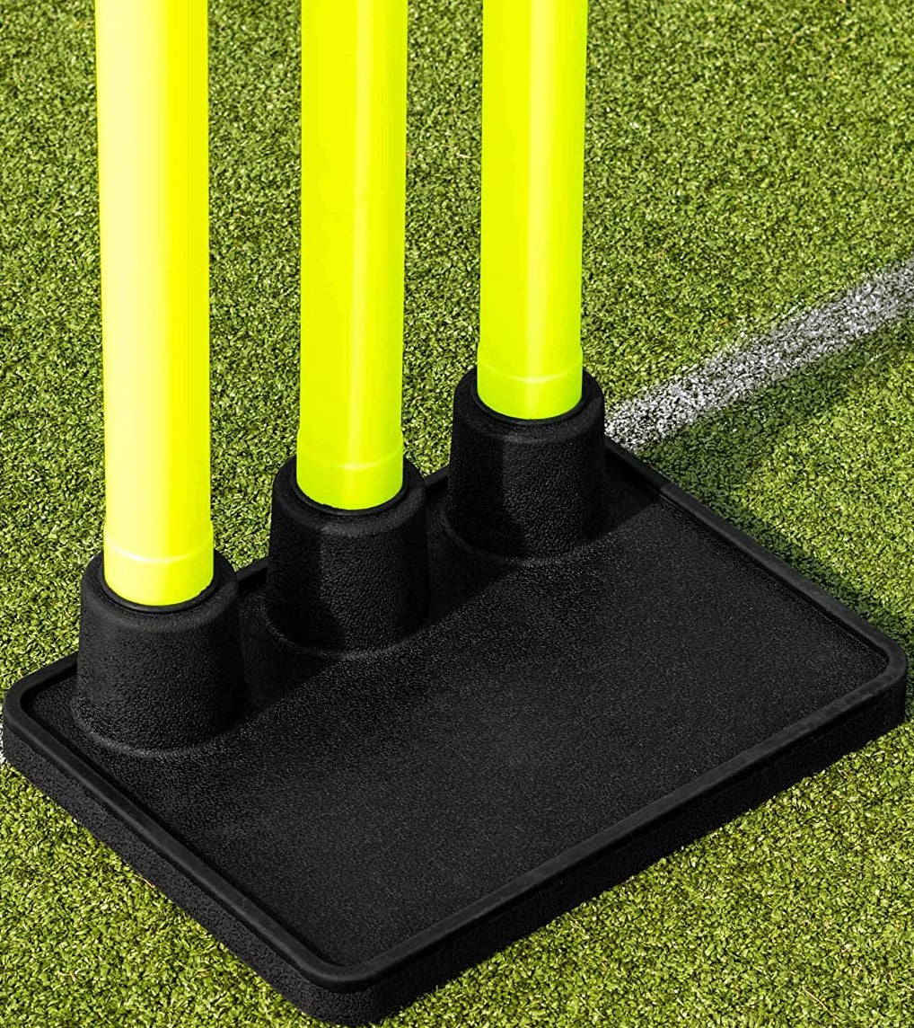 Three yellow cricket stumps stand upright in the Martin Berrill Sports Heavy Duty Rubber Flexi Stump Base (Black) on green artificial turf, with a white line visible in the background.