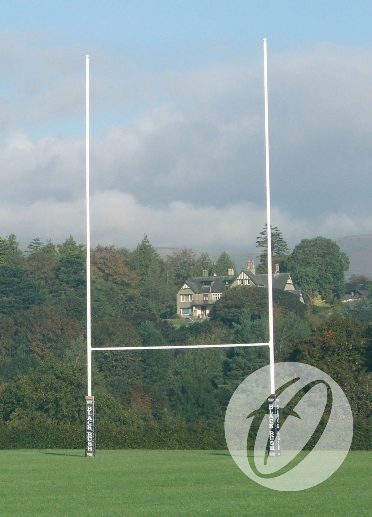 A set of Harrod Sport No. 2 Steel Rugby Posts - 9M Socketed stands on a grassy field with tall trees and a large house in the background under a partly cloudy sky.
