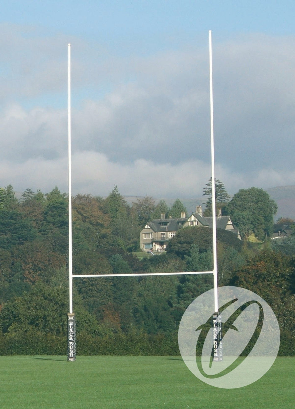 A set of Harrod Sport No. 2 Steel Rugby Posts - 9M Socketed stands on a grassy field with tall trees and a large house in the background under a partly cloudy sky.