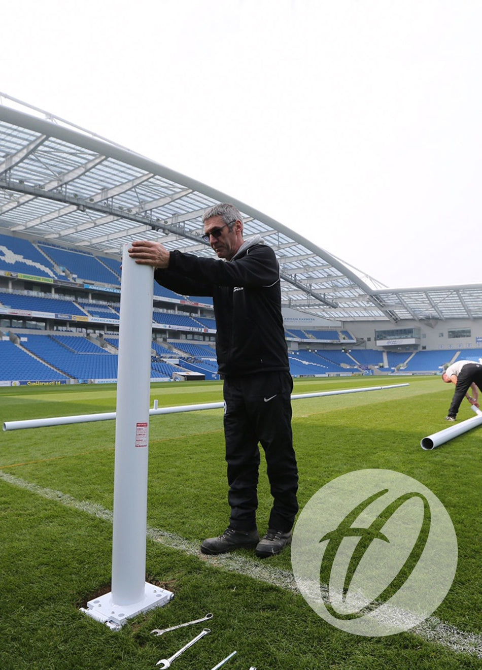 A man assembles Harrod Sport Millennium Rugby Posts - 17M Hinged on a sports field, with tools on the grass nearby. The stadium features blue seats, a partial roof, and another person working in the background.