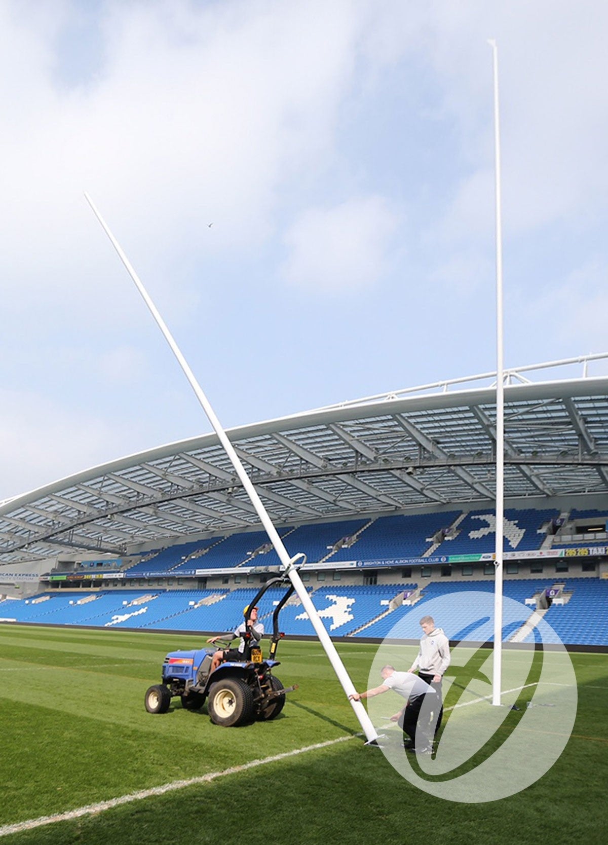 Two people use a small tractor to install Harrod Sport Millennium Rugby Posts - 17M Hinged on a stadium field, with blue seats and a partially covered roof in the background.