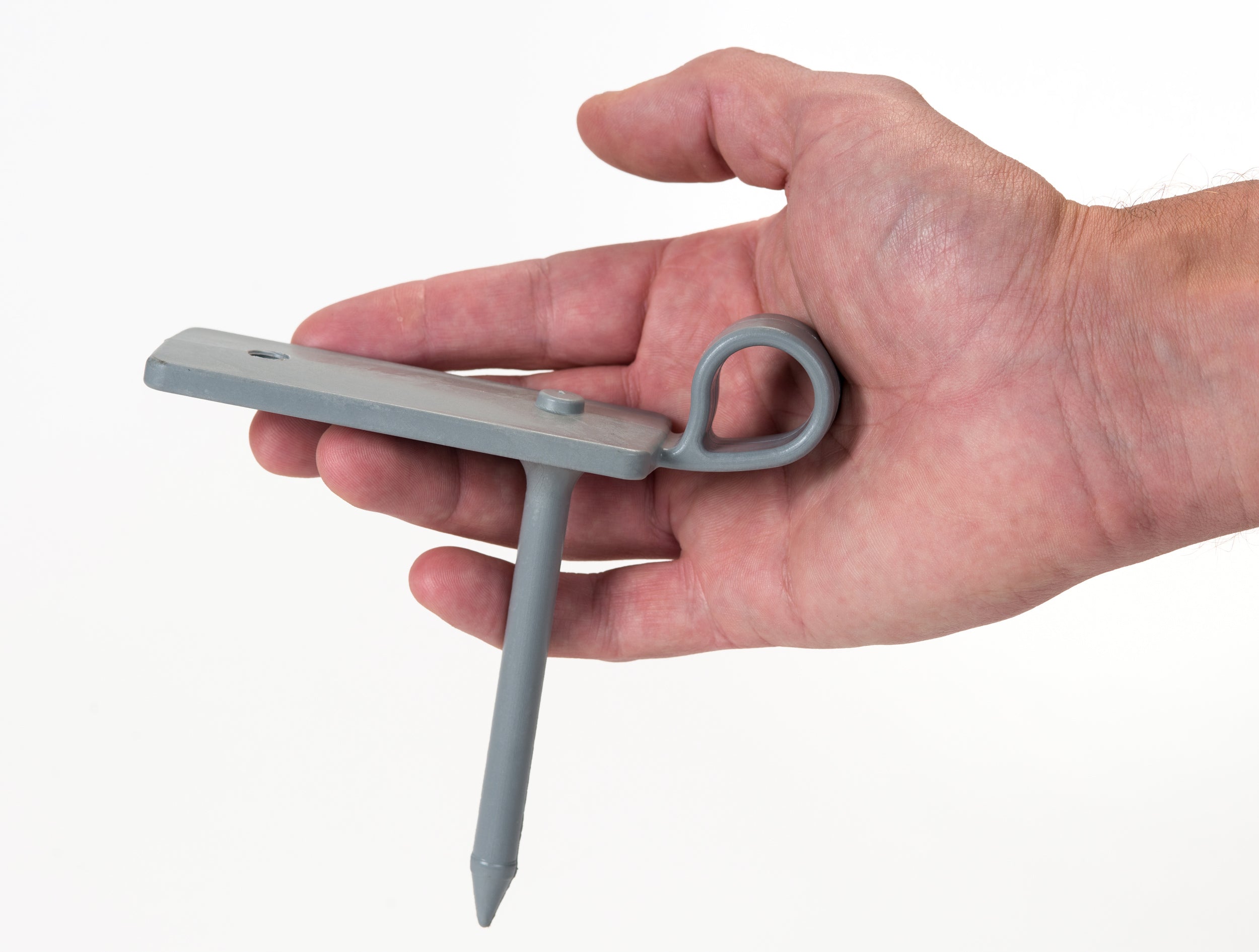 A hand holds the Fusion Multi-stump Spike by Fusion—a gray metal cricket accessory with a flat plate, circular loop, and long pointed stump spike—designed for hard ground, against a plain white background.