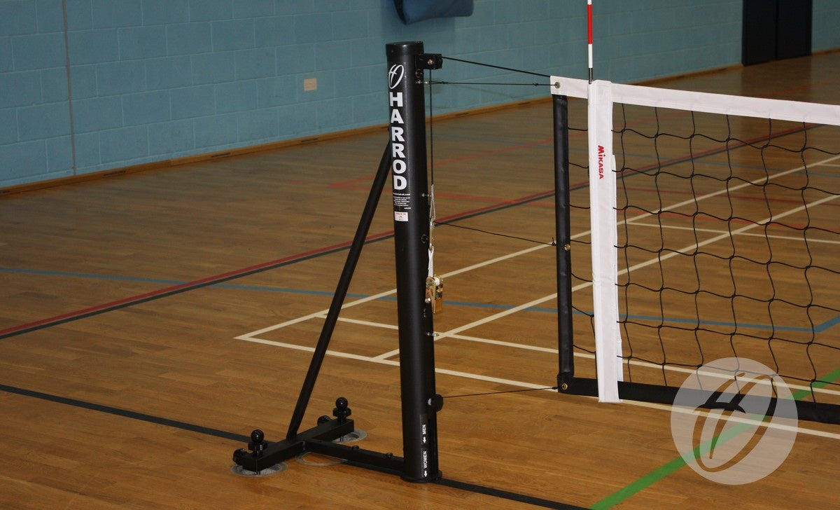 A close-up of Harrod Sport Sitting Volleyball Posts with a steel slider supports a volleyball net on a wooden gym floor, featuring colored court lines and a blue wall in the background.
