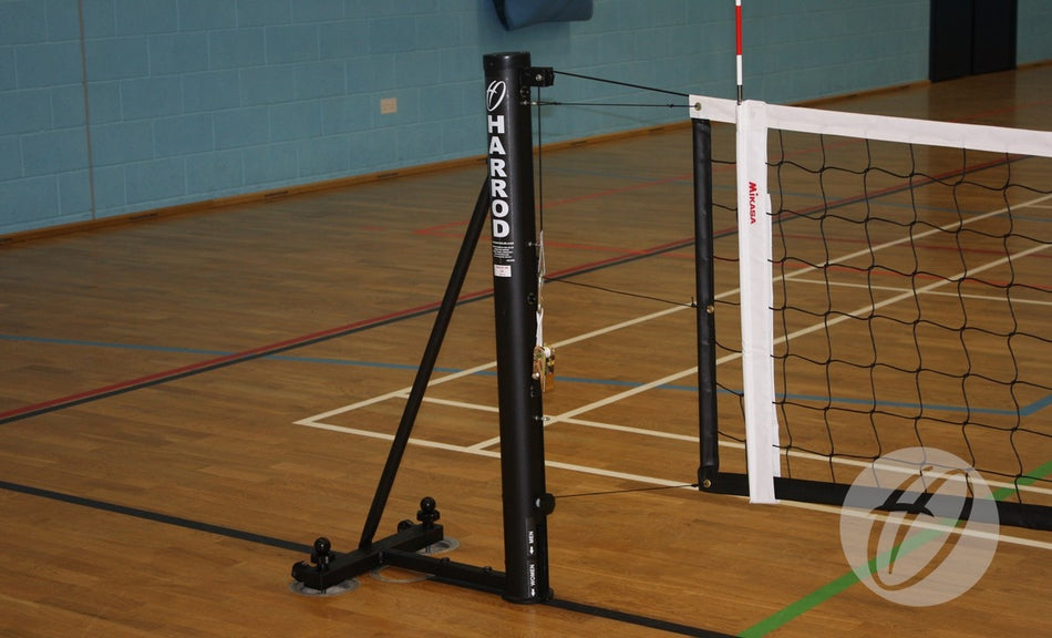 A close-up of Harrod Sport Sitting Volleyball Posts with a steel slider supports a volleyball net on a wooden gym floor, featuring colored court lines and a blue wall in the background.