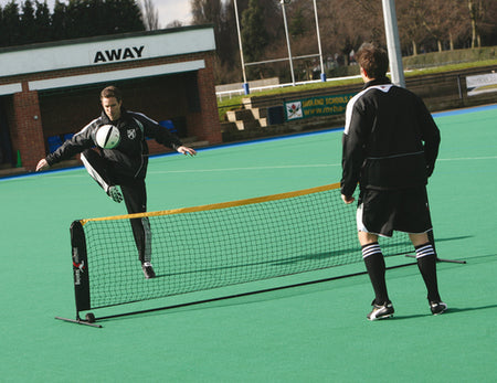 Two men play head tennis outdoors using the Precision Training Precision Soccer Skills Net (single); one leaps to kick the ball over the net while the other stands ready. Both wear black athletic outfits and striped socks.