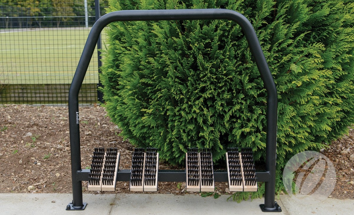 A Harrod Sport Socketed Angle Boot Wipers unit with black metal frame and beech timber brushes is installed outdoors on concrete, positioned in front of green bushes and a wire fence.