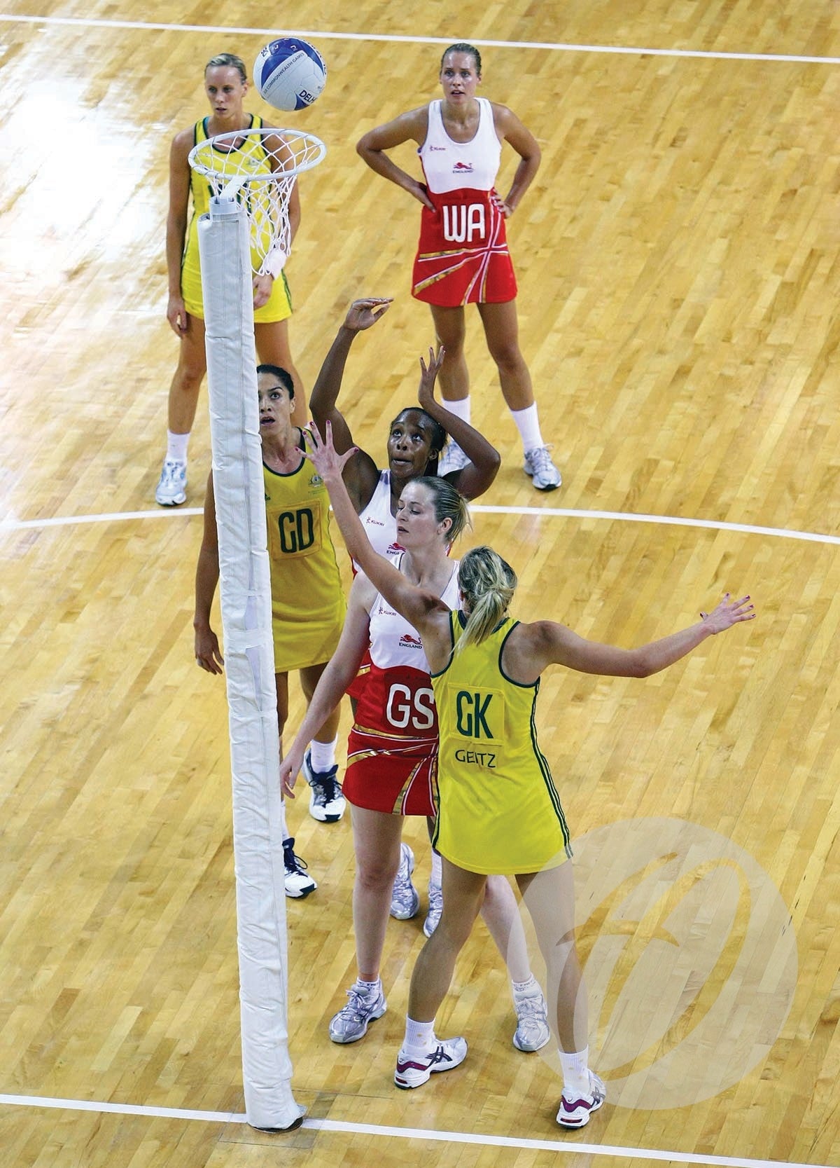 A women’s netball game on a wooden court shows yellow players defending red as the ball nears Harrod Sport Sprung Floor Socketed International Netball Posts. Four players are near the posts, with two others standing behind them.