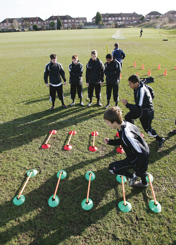 A group of boys in sportswear watch two others navigate agility poles and the Precision Speed Agility Cone Set by Precision Training on a grassy field, with houses and trees visible in the background.