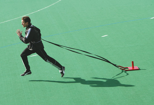 Wearing sportswear, a person sprints across a green field with the Precision Training Precision Speed Sled strapped to their waist, using resisted sprinting to enhance power and speed.