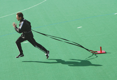 Wearing sportswear, a person sprints across a green field with the Precision Training Precision Speed Sled strapped to their waist, using resisted sprinting to enhance power and speed.