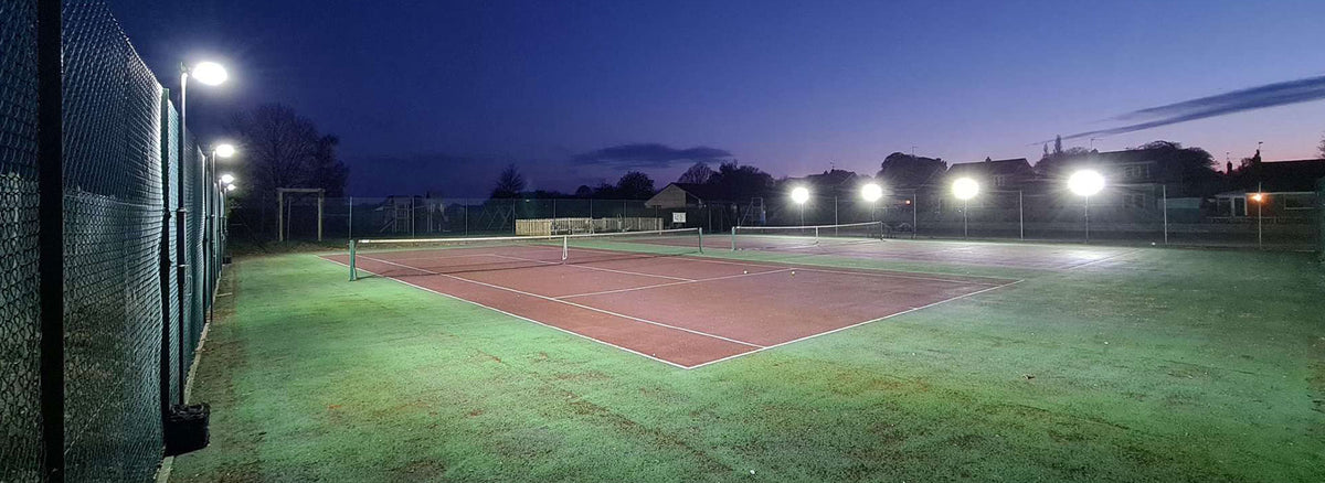 A tennis court with sports fencing is brightly lit at dusk using the Ritelite Sports Lite Fence Mast Adapter, set against a clear deep blue sky with silhouettes of trees and buildings in the background.
