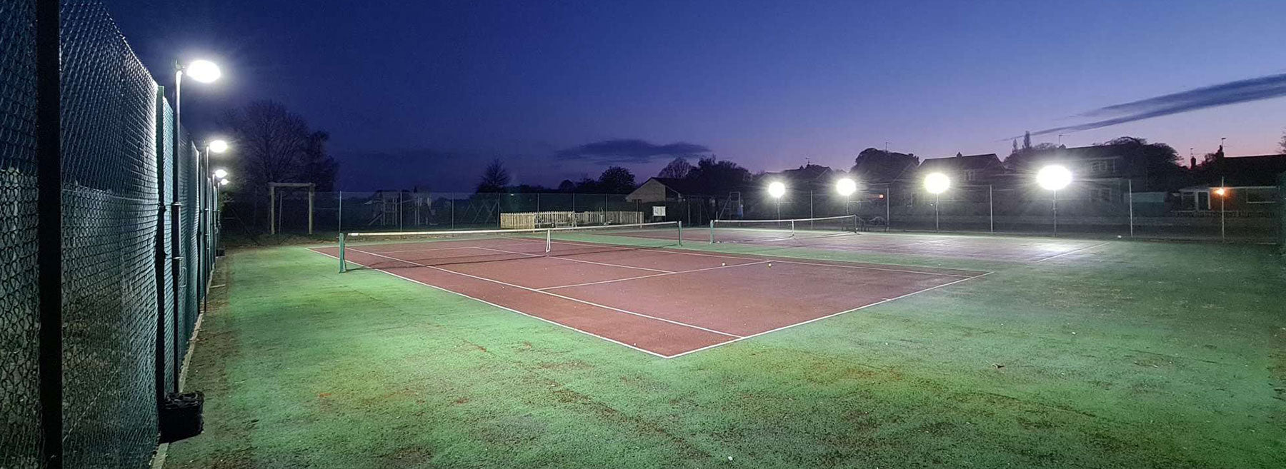 A tennis court with sports fencing is brightly lit at dusk using the Ritelite Sports Lite Fence Mast Adapter, set against a clear deep blue sky with silhouettes of trees and buildings in the background.