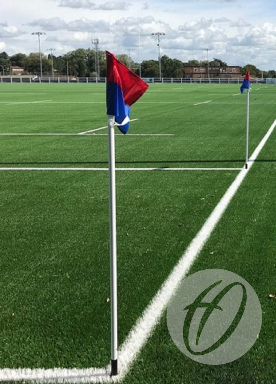 A sports field features artificial green turf, white boundary lines, and Harrod Sport 32mm Spring Back Corner Pole & Socket with red and blue flags. Buildings and trees appear in the background under a partly cloudy sky.