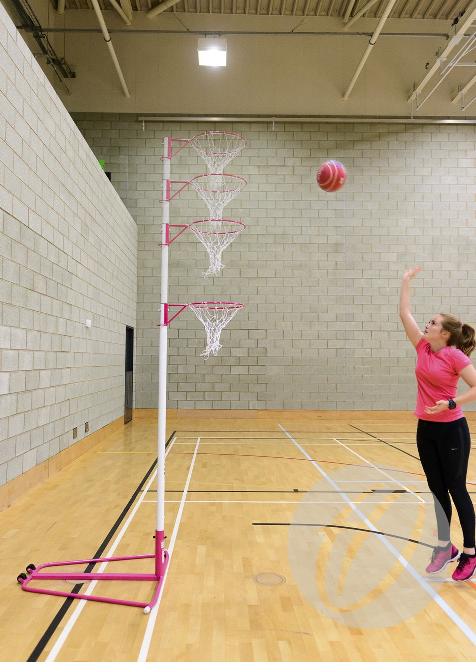 A woman in pink sportswear shoots a pink basketball toward a tall Harrod Sport Single Telescopic Netball Post in a gymnasium with wooden floors and brick walls.