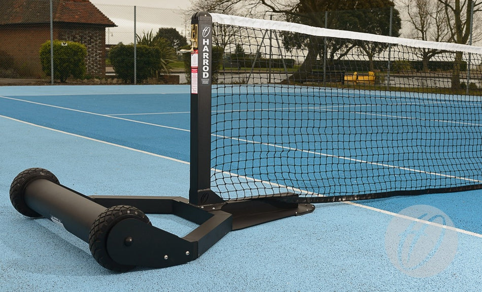 A close-up of Harrod Sport's Harrod UK Integral Weighted Tennis Posts with wheels on a blue outdoor court, the net partially visible and houses, trees, and a fence in the background.