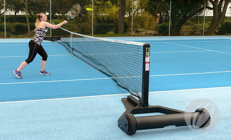A woman in athletic wear hits a tennis ball over the net on a blue outdoor court. The net is supported by Harrod Sport Harrod UK Integral Weighted Tennis Posts with black wheeled bases. Trees and a fence appear in the background.