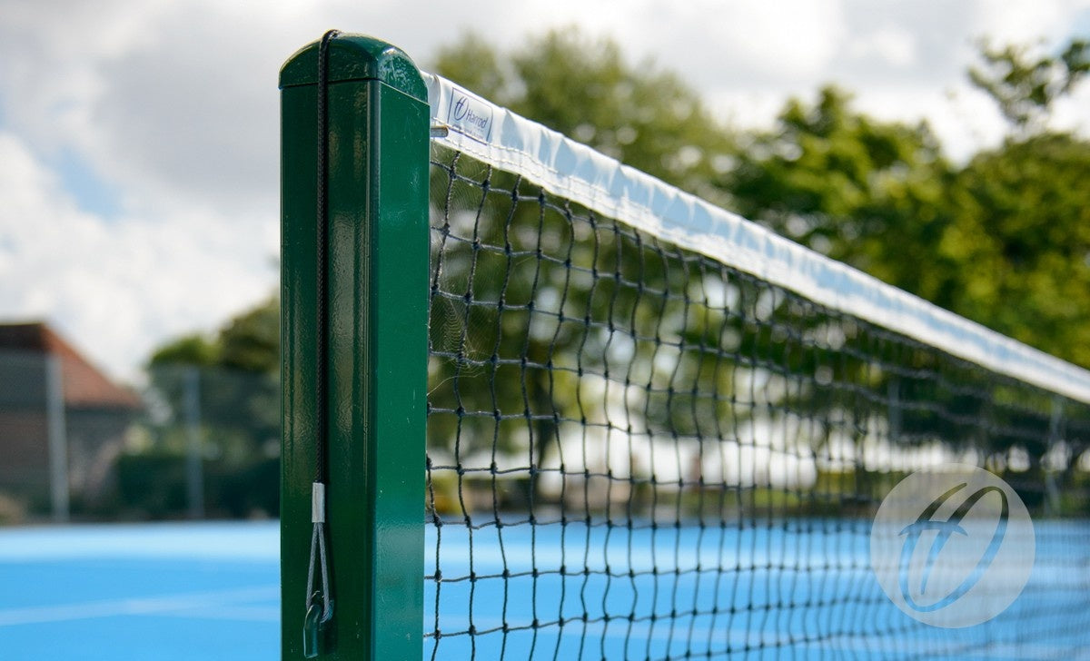 Close-up of Harrod Sport S8 Tennis Posts C/W Locking Sockets, powder coated green, installed on an outdoor blue court. The net and post are sharp against a blurred background of trees and cloudy sky.