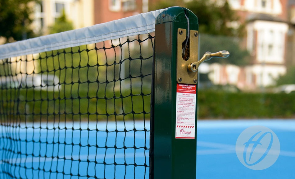 Close-up of Harrod Sport S8 Tennis Posts C/W Locking Sockets with a powder-coated green finish and metal winding mechanism, on a blue outdoor court. Red information sticker visible; blurred buildings and greenery in the background.