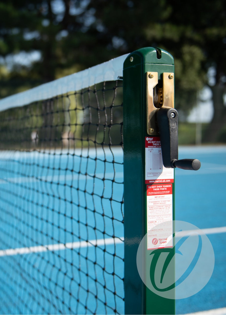 Close-up of the Harrod Sport Tennis Torque Limiter featuring a black handle and adjustment mechanism on a bright blue outdoor tennis court, with trees and greenery blurred in the background.