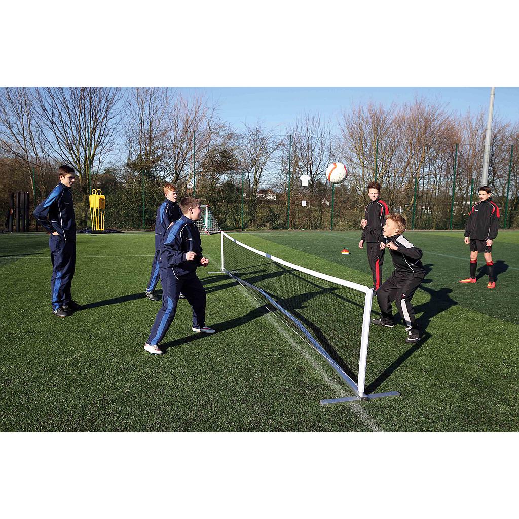 Five boys play a game on grass, using the Precision Training Compact Tennis Net Set and heading a ball. Two boys in tracksuits, resembling tennis coaches, observe nearby. Trees and sports gear are visible in the background.