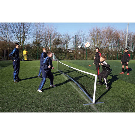 Five boys play a game on grass, using the Precision Training Compact Tennis Net Set and heading a ball. Two boys in tracksuits, resembling tennis coaches, observe nearby. Trees and sports gear are visible in the background.