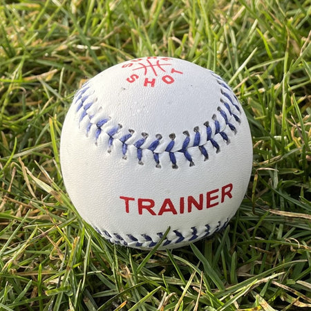 The Sure Shot Trainer Rounders Ball by Sure Shot is a white, durable training ball with a PVC leather cover, blue stitching, and red "TRAINER" text. It also features red text and a partial logo on the top, shown resting on green grass.