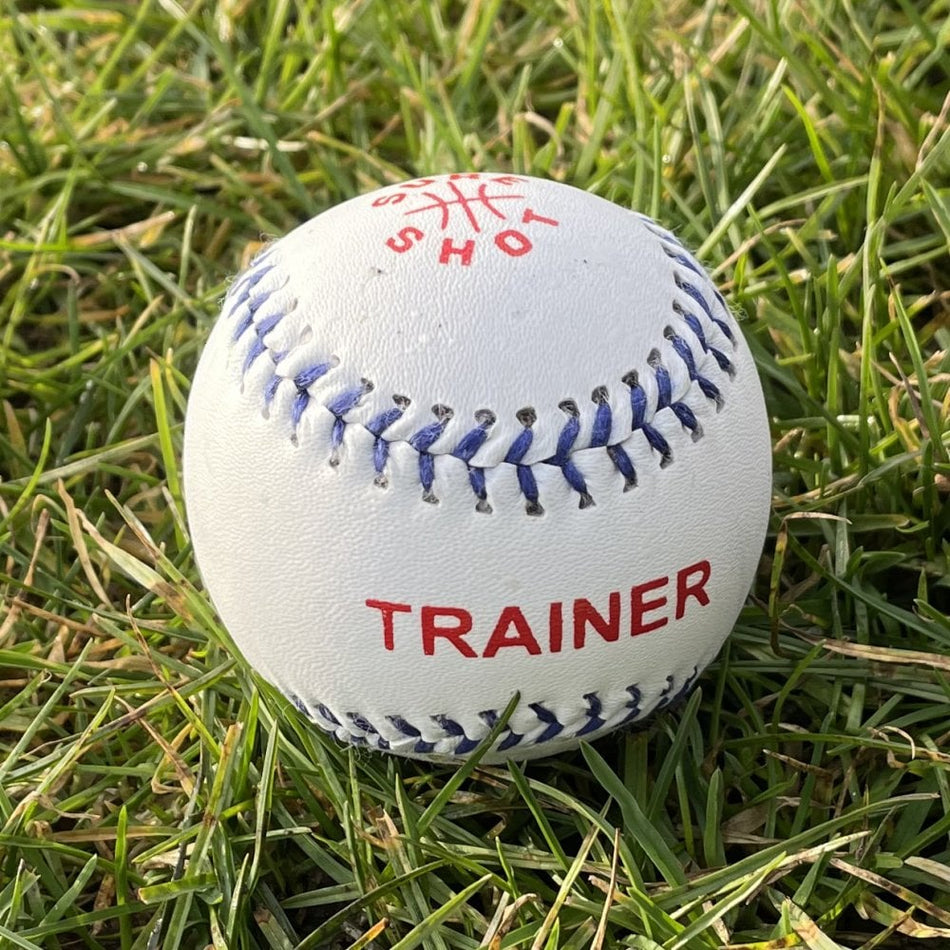 The Sure Shot Trainer Rounders Ball by Sure Shot is a white, durable training ball with a PVC leather cover, blue stitching, and red "TRAINER" text. It also features red text and a partial logo on the top, shown resting on green grass.