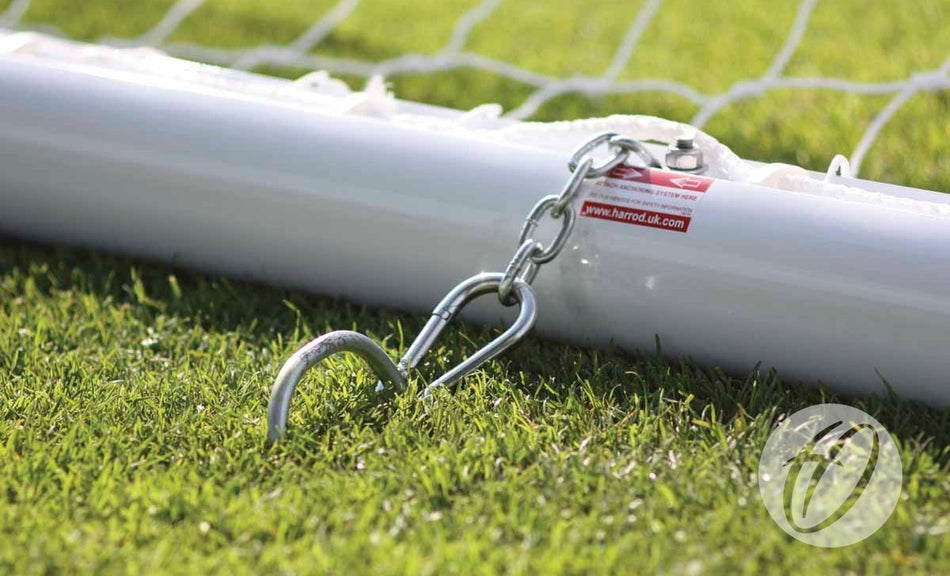 A close-up of a white soccer goalpost anchored to grass with Harrod Sport U Peg Anchors (10MM x 450MM) and a metal chain for stability. The netting is firmly secured to the goalpost.