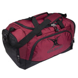 The Gilbert V3 Club Player Holdall is a maroon sports bag with multiple zippered compartments, black straps and handles, a padded shoulder strap for comfort, and features the Gilbert logo on the side pocket.