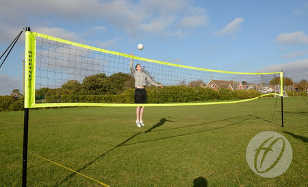 A person jumps to hit a volleyball over the Harrod Sport Portable Volleyball set on a grassy field, with houses, trees, and a blue sky in the background.