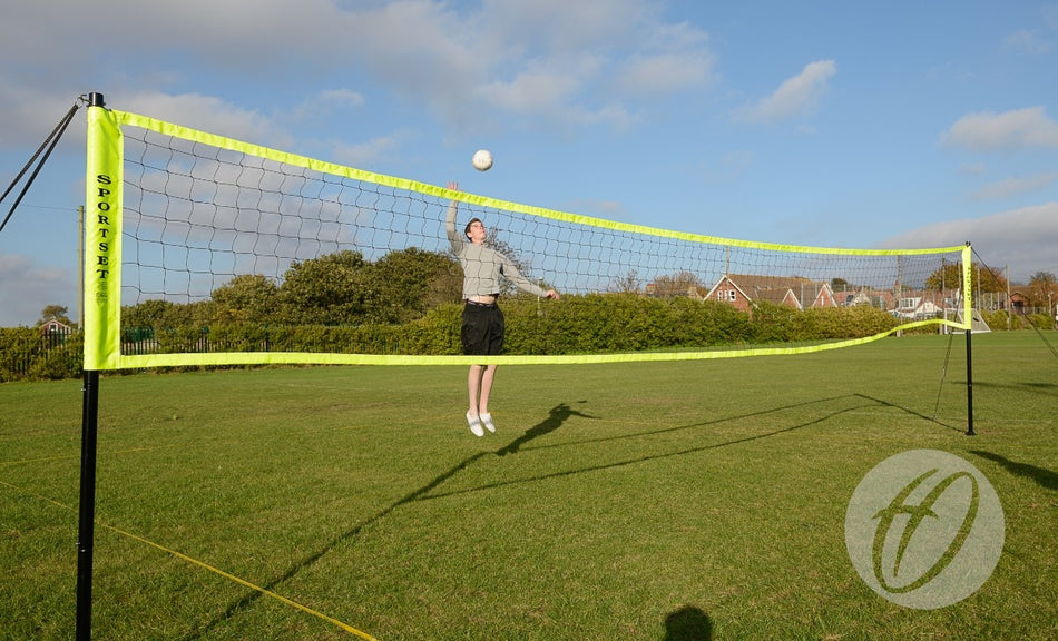 A person jumps to hit a volleyball over the Harrod Sport Portable Volleyball set on a grassy field, with houses, trees, and a blue sky in the background.