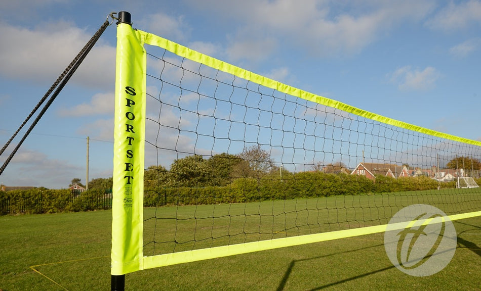 A Harrod Sport Portable Volleyball set with yellow borders is assembled on grass, with houses, trees, and a blue sky with scattered clouds in the background.