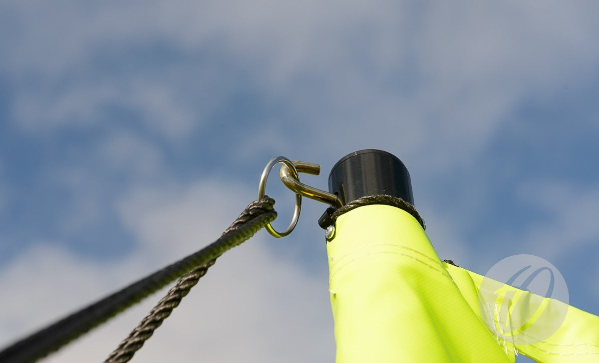 Close-up of a black rope and metal hook attaching yellow fabric on the Harrod Sport Portable Volleyball net, set against a blue sky with clouds. The image highlights the secure connection point of rope, hook, and fabric.