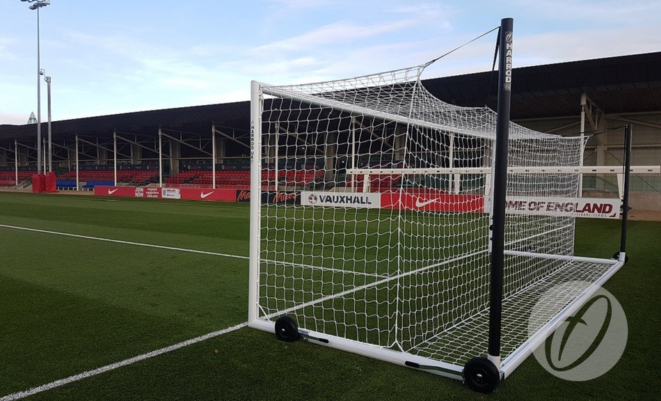 A Harrod Sport Weighted UEFA Portagoal with high raise wheels stands on a green artificial turf field, near a stadium with red seats and a Home of England sign in the background.