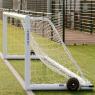 A black and white soccer ball rests against the net inside a Harrod Sport 4G Weighted Portagoal - 5v5 (4.88 x 1.22m) on bright green artificial turf, its reinforced aluminium frame standing out. In the background, a metal fence and tennis courts emphasize the convenience and portability of this premium football goal.