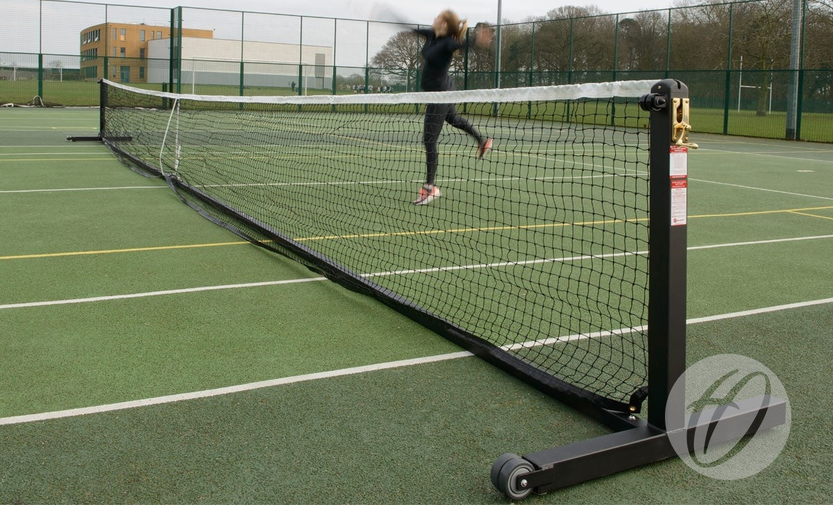 A person jumps to hit a tennis ball on an outdoor court equipped with the Harrod Sport Wheelaway Freestanding Tennis Post, featuring wheeled posts and a heavy duty brass winder. Trees, a building, and fencing appear in the background.