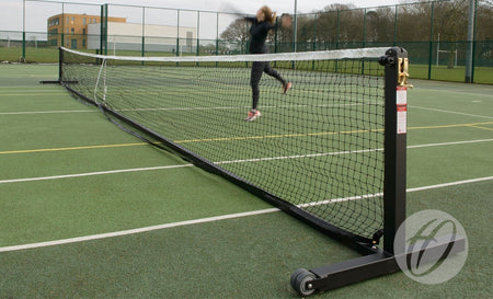 A person jumps to hit a tennis ball on an outdoor court equipped with the Harrod Sport Wheelaway Freestanding Tennis Post, featuring wheeled posts and a heavy duty brass winder. Trees, a building, and fencing appear in the background.
