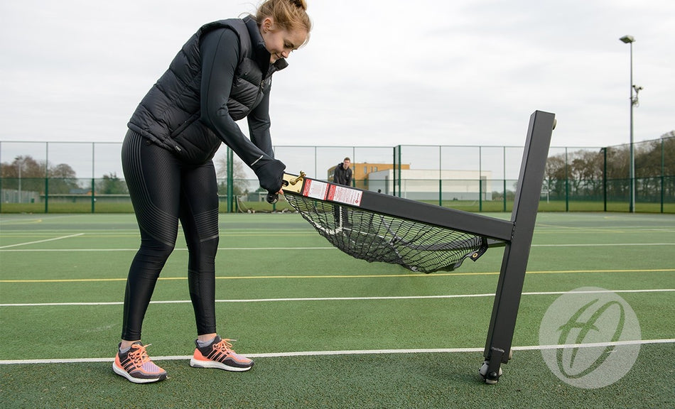 A woman in athletic wear assembles a Harrod Sport Wheelaway Freestanding Tennis Post with steel posts for a portable net on an outdoor court. The partially assembled frame is angled, while another person stands by the fence in the background.