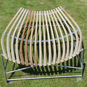 The Wooden Slip Catch Cradle by Martin Berrill Sports features evenly spaced curved hardwood ash slats on a metal frame, resembling a cricket fielding aid, and is shown from above resting on green grass.