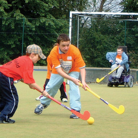 Two boys play field hockey outdoors with Eurohoc Floorball Zone Hockey Set sticks, while another boy in a wheelchair watches. All wear casual clothes and enjoy a game of Eurohoc Zone Hockey under clear skies.